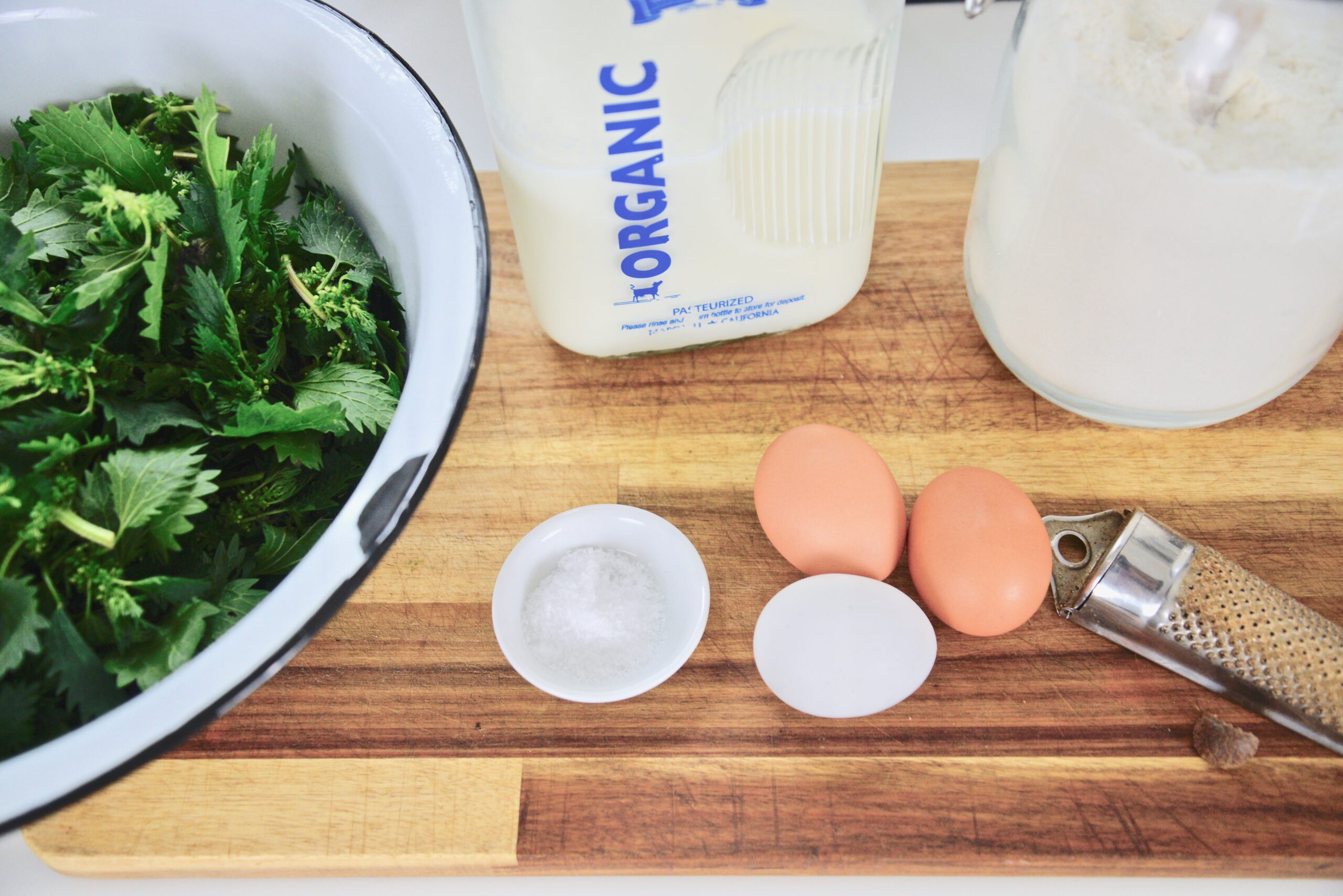 fresh stinging nettles in bowl, milk, flour, eggs, salt, and nutmeg in nutmeg grater